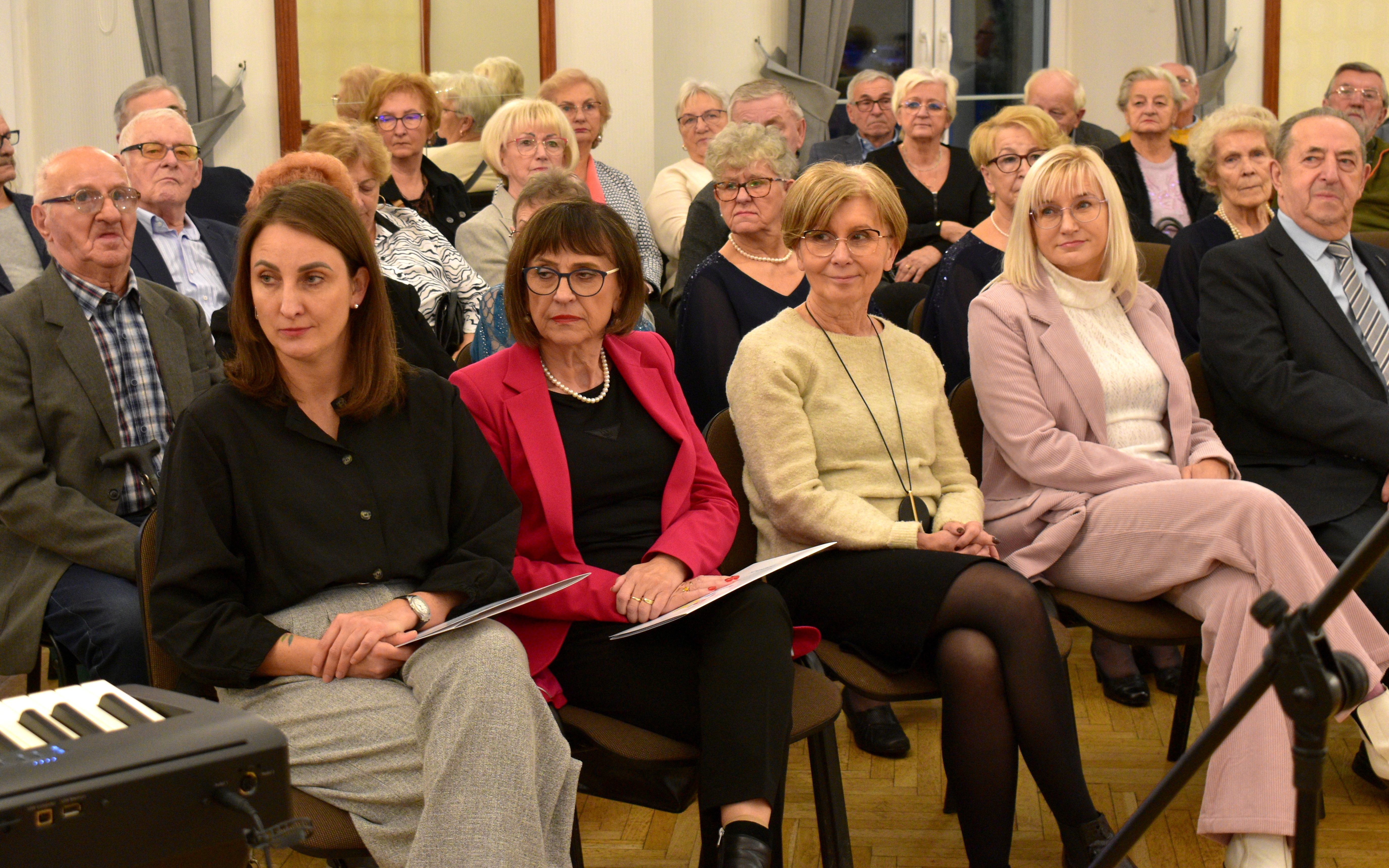 Widownia złożona gł&oacute;wnie z&nbsp;os&oacute;b starszych siedzących w&nbsp;rzędach na&nbsp;krzesłach w&nbsp;eleganckiej sali. Na pierwszym planie cztery kobiety w&nbsp;odświętnych strojach. W tle widać inne osoby siedzące i&nbsp;wsłuchujące się w&nbsp;wydarzenie.