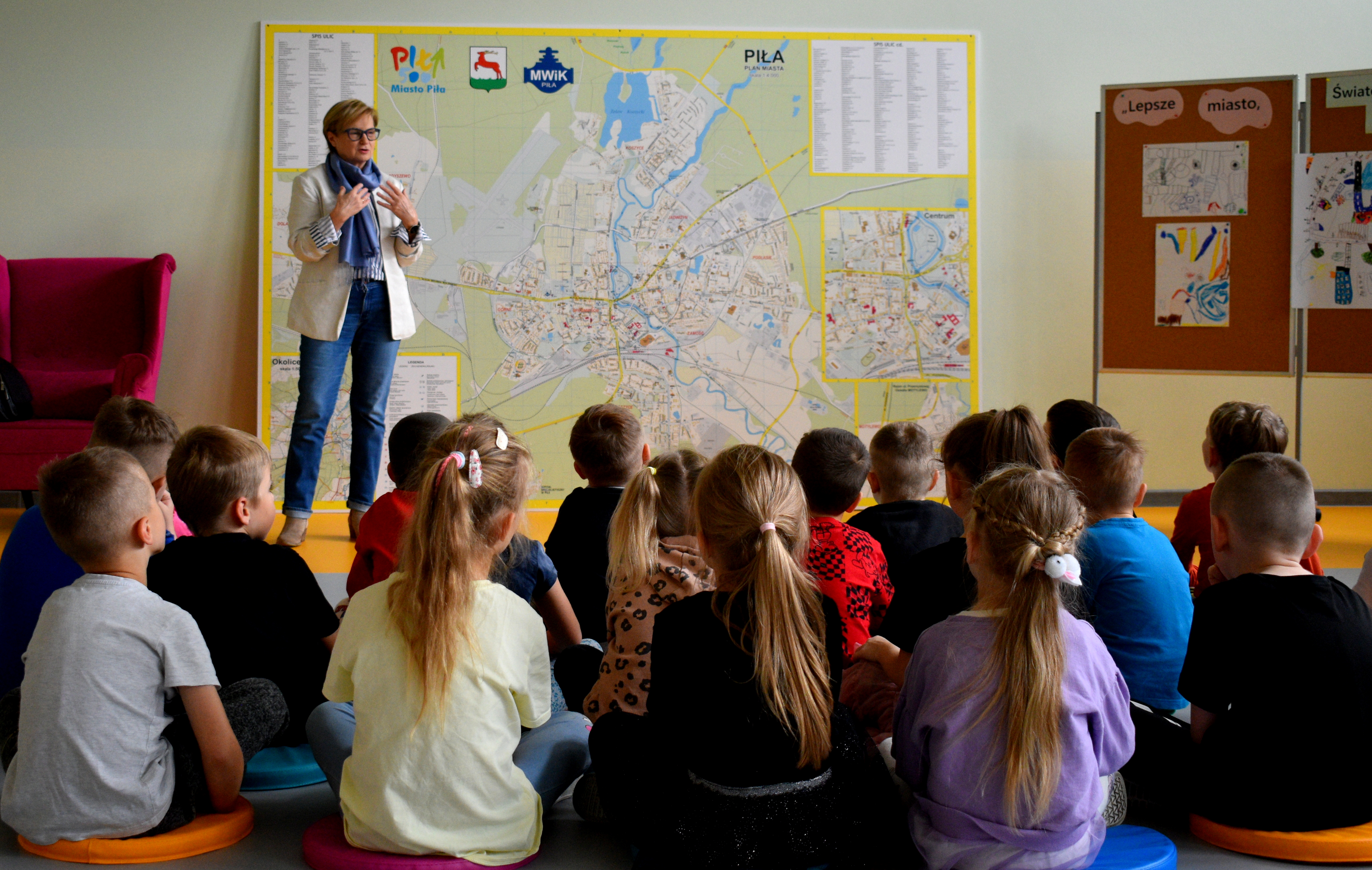 Na zdjęciu widać kobietę stojącą przed dużą mapą miasta Piła, kt&oacute;ra prezentuje coś grupie dzieci siedzących przed nią na&nbsp;poduszkach. Mapa zajmuje całą szerokość ściany za nią i&nbsp;przedstawia topografię miasta, oznaczenia dr&oacute;g i&nbsp;obszary miejskie. Po prawej stronie widoczne są tablice z&nbsp;dziecięcymi rysunkami i&nbsp;napisami &bdquo;Lepsze miasto, Światowy Dzień Miast&rdquo;.