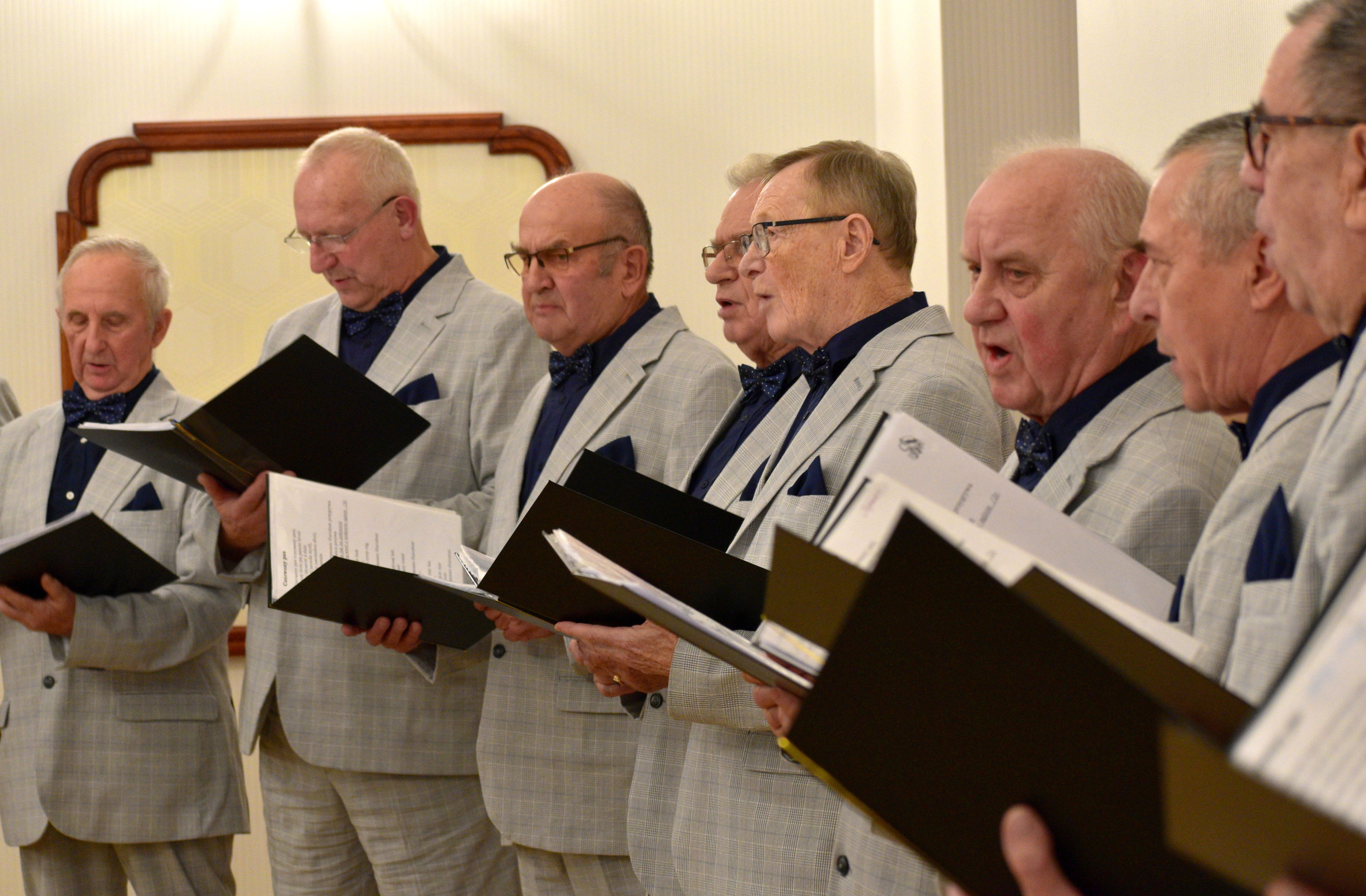 Ch&oacute;r męski w&nbsp;jednolitych, jasnoszarych garniturach z&nbsp;ciemnymi koszulami i&nbsp;muszkami wykonuje utw&oacute;r muzyczny. Mężczyźni stoją w&nbsp;p&oacute;łkolu, trzymając czarne teczki z&nbsp;nutami. 