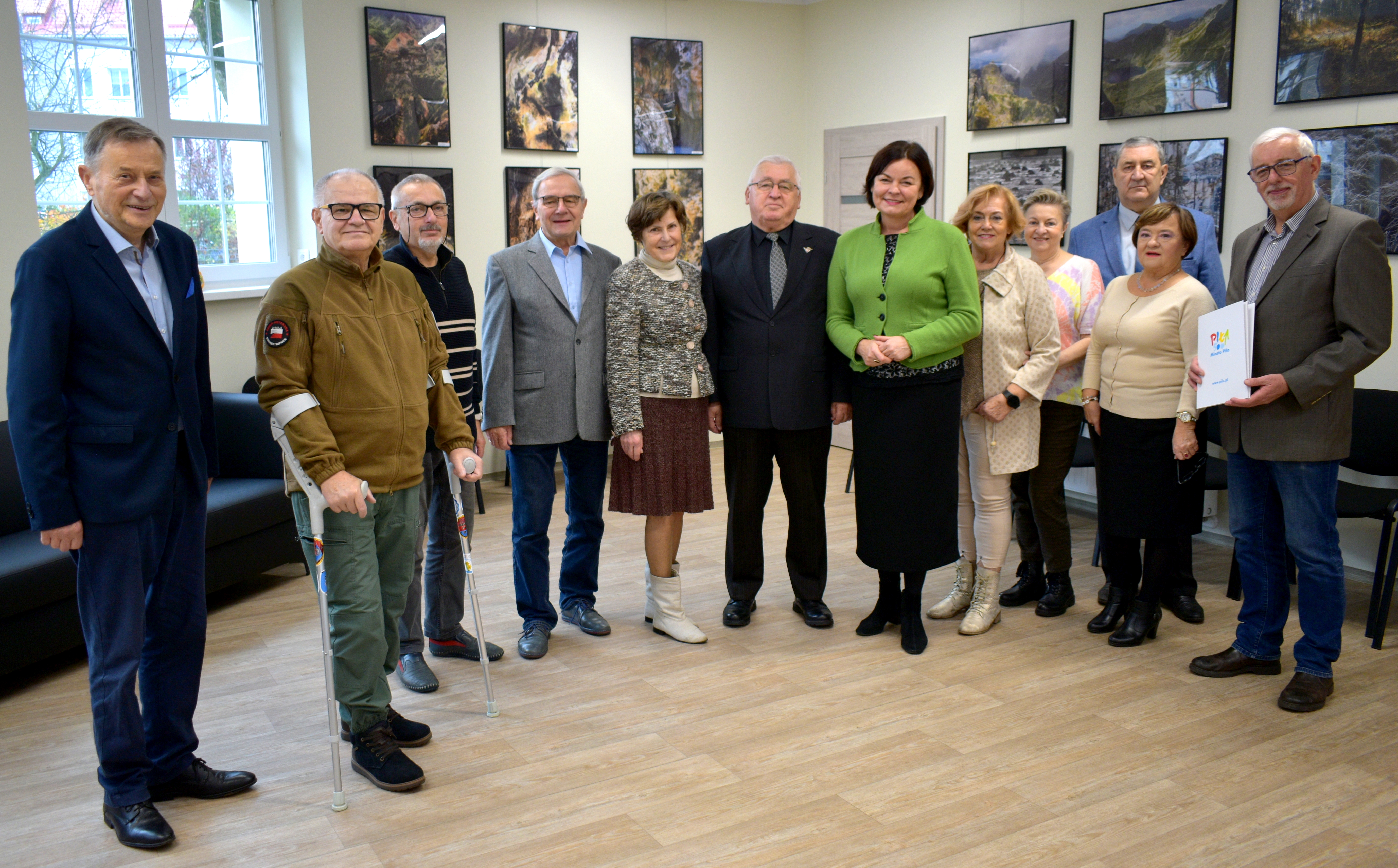 Grupa dwunastu os&oacute;b pozujących wsp&oacute;lnie do zdjęcia w&nbsp;jasnym pomieszczeniu. Na pierwszym planie stoją osoby w&nbsp;r&oacute;żnym wieku, ubrane elegancko. Wśr&oacute;d nich znajdują się mężczyzna z&nbsp;kulami ortopedycznymi oraz&nbsp;kobieta w&nbsp;zielonym żakiecie. Po prawej stronie mężczyzna w&nbsp;marynarce trzyma teczkę z&nbsp;logo &bdquo;Miasto Piła&rdquo;. W tle widać rząd zdjęć krajobrazowych zawieszonych na&nbsp;ścianie oraz&nbsp;dużą sofę przy oknie. 