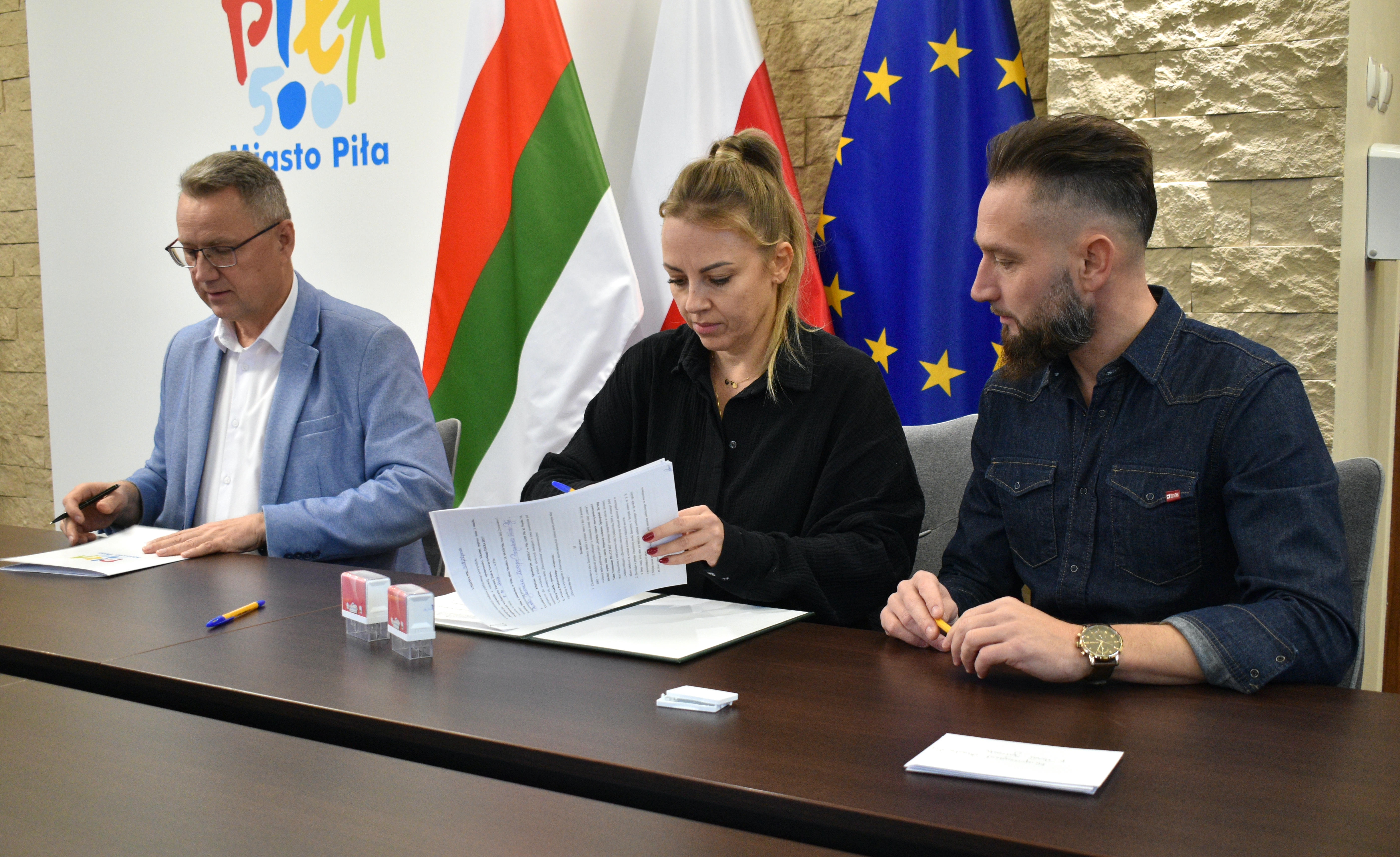 Kobieta i&nbsp;dw&oacute;ch mężczyzn podpisują umowy w&nbsp;pomieszczeniu, w&nbsp;tle flaga Piły, Polski i&nbsp;Unii Europejskiej