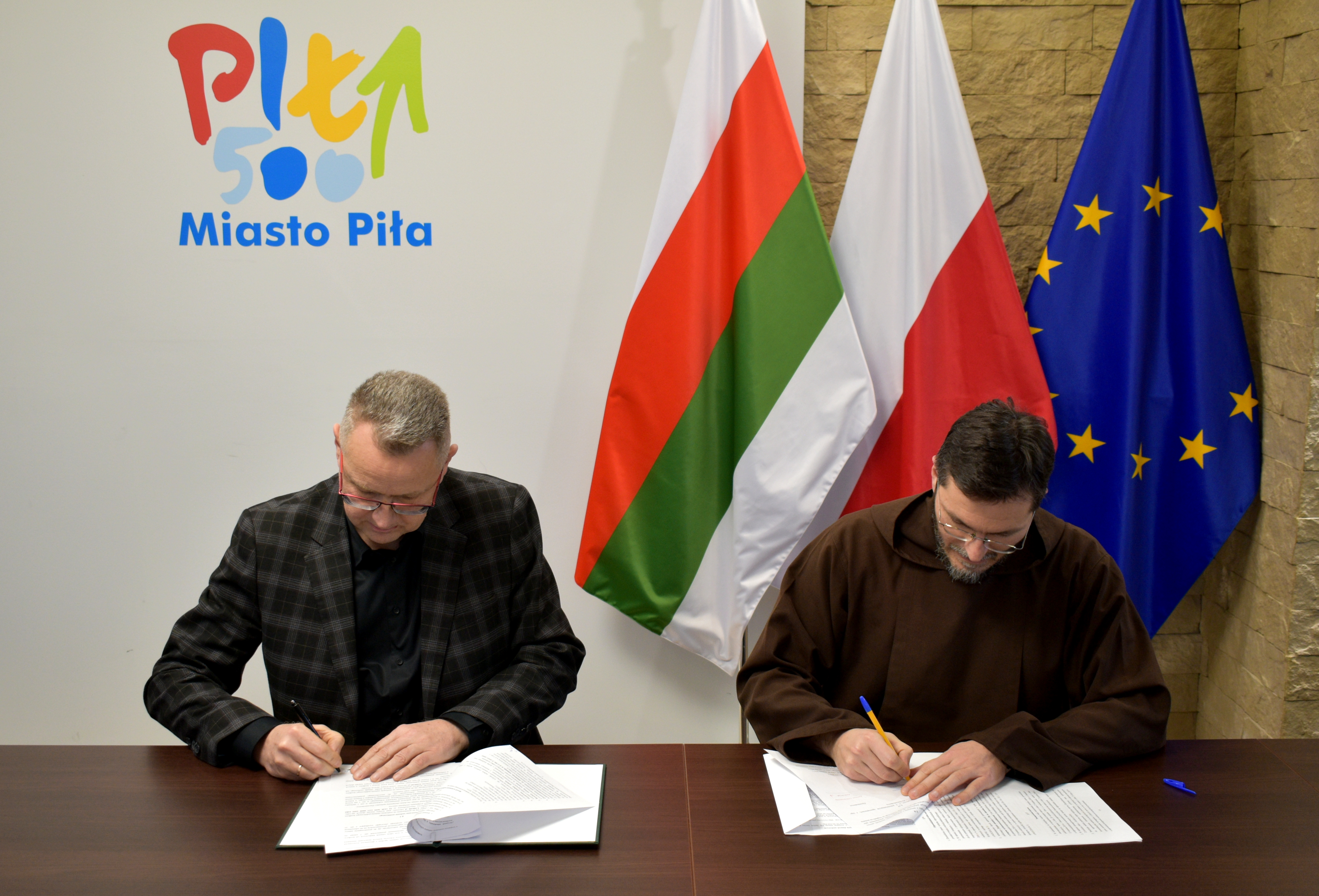 Zastępca prezydenta miasta Piły podpisuje dokumenty razem z&nbsp;przedstawicielem parafii Rzymskokatolickiej pw. św. Antoniego w&nbsp;Pile. Obaj mężczyźni siedzą przy stole, na&nbsp;kt&oacute;rym leżą dokumenty i&nbsp;mają w&nbsp;rękach długopisy. W tle widoczne są flagi Polski, Unii Europejskiej oraz&nbsp; miasta Piły.