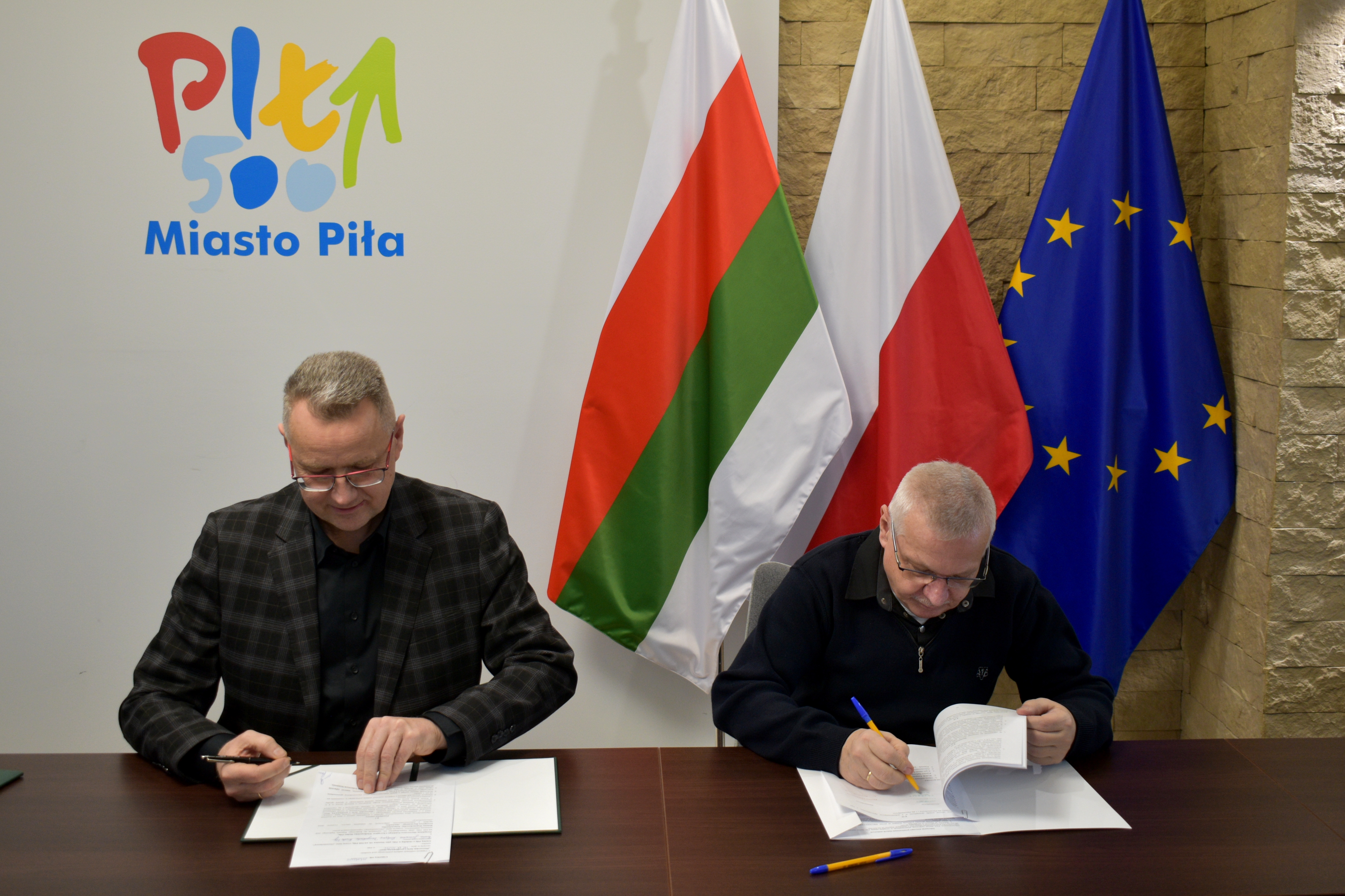 Zastępca prezydenta miasta Piły podpisuje dokumenty z&nbsp;przedstawicielem Związku Harcerstwa Polskiego Chorągiew Wielkopolska Hufiec Piła. Obaj mężczyźni siedzą przy stole, na&nbsp;kt&oacute;rym leżą dokumenty i&nbsp;mają w&nbsp;rękach długopisy. W tle widoczne są flagi Polski, Unii Europejskiej oraz&nbsp; miasta Piły.