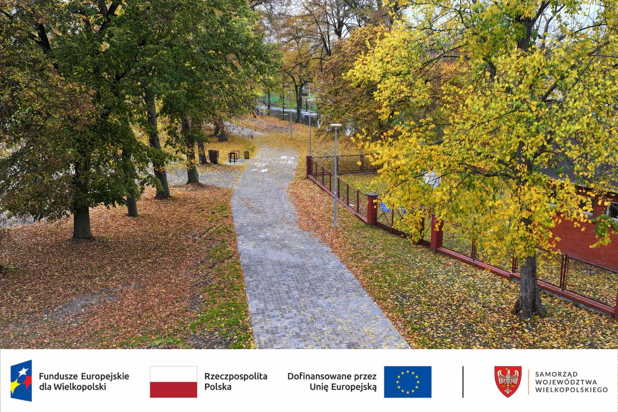 Jesienny skwer z&nbsp;nowym ciągiem pieszo-rowerowym wśr&oacute;d drzew o&nbsp;ż&oacute;łtych i&nbsp;zielonych liściach. U dołu zdjęcia znajdują się logotypy Funduszy Europejskich dla Wielkopolski, Rzeczypospolitej Polskiej, Unii Europejskiej oraz&nbsp;Samorządu Wojew&oacute;dztwa Wielkopolskiego.