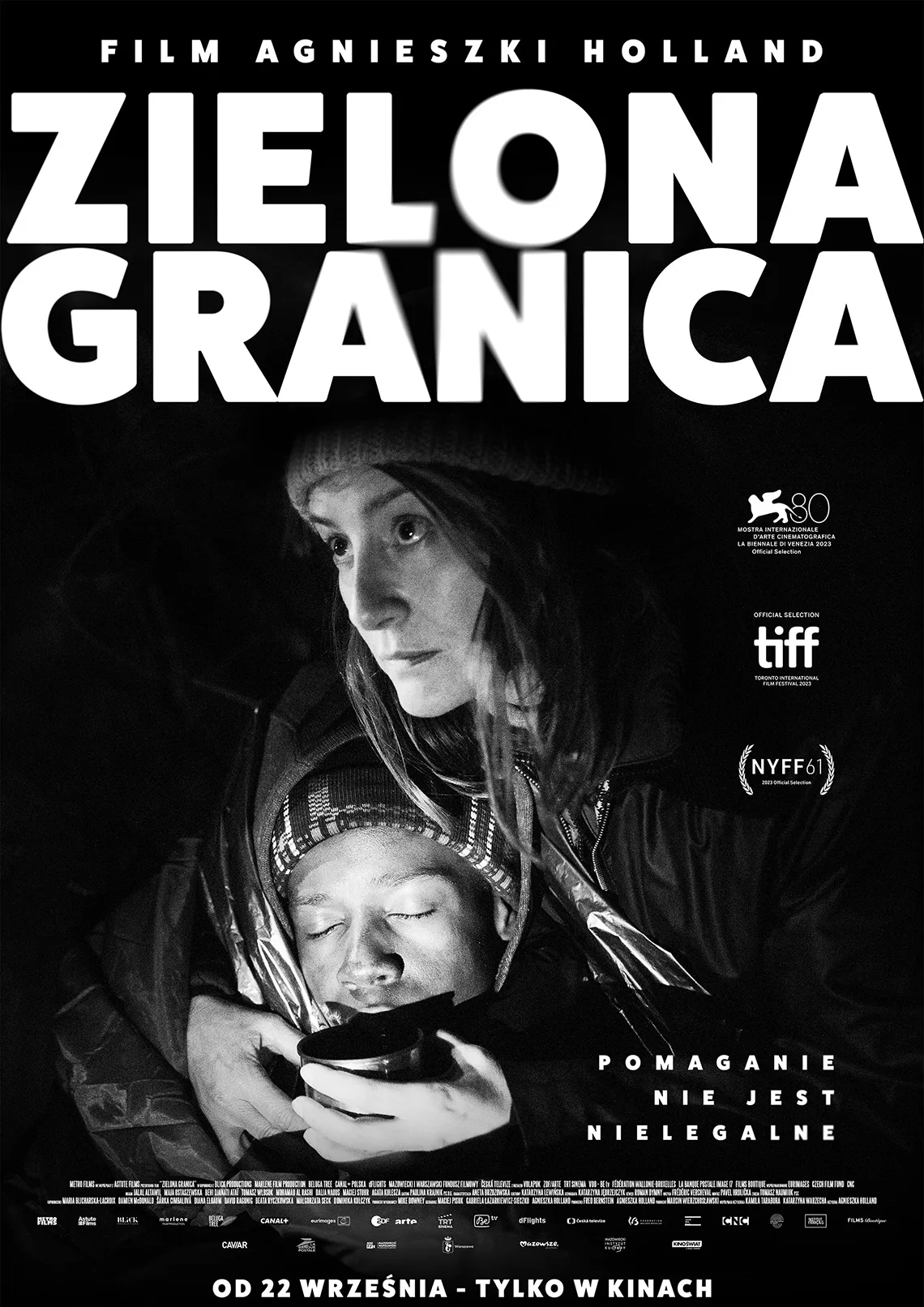 Czarno białe zdjęcie kobiety, przy niej mężczyzna z&nbsp;zamkniętymi oczami. Treść: Film Agnieszki Holland Zielona Granica, pomaganie nie jest nielegalne.