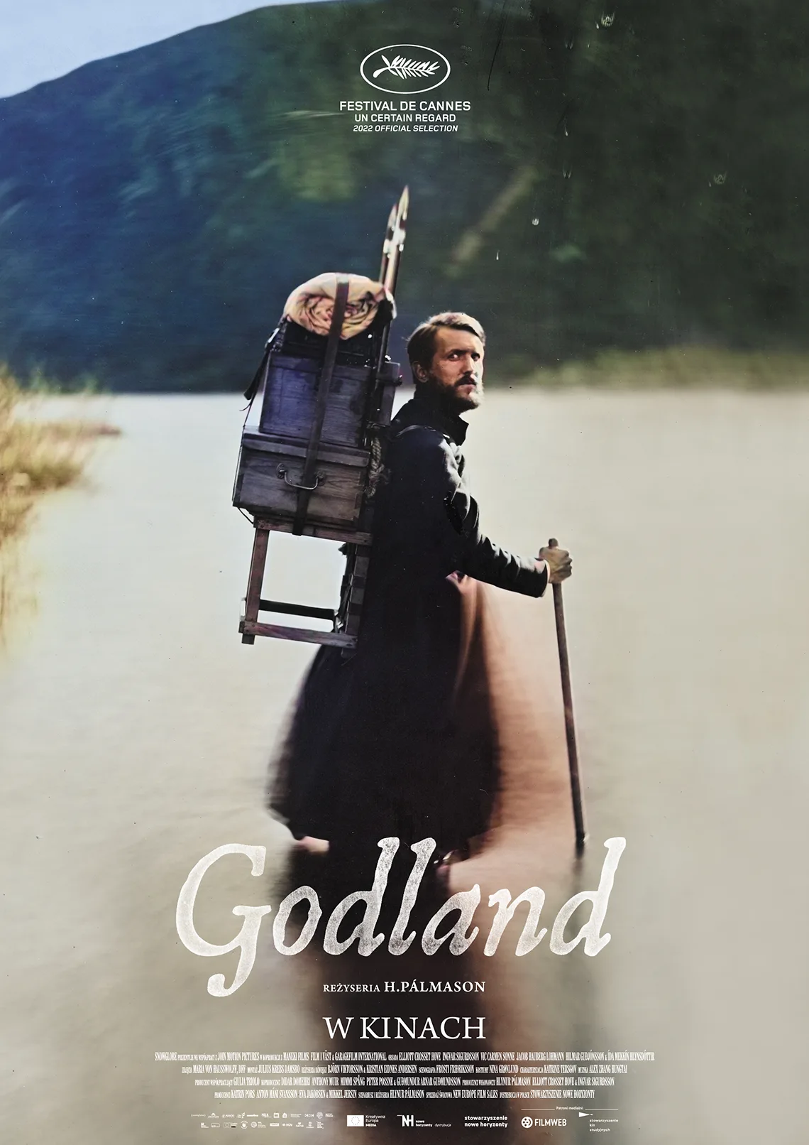 WDK w RCK: Godland Mężczyzna z bagażami na placach, podpiera się kijem i ogląda za siebie. W tle zieleń. Treść: Godland.
