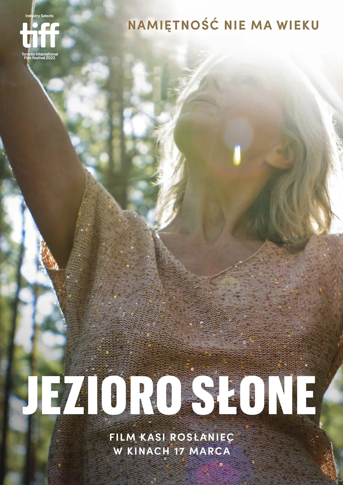 PLAKAT. Portret kobiety stojącej pomiędzy drzewami, nad jej głową promienie słoneczne. Treść: Namiętność nie ma wieku, jezioro słone. Film Kasi Rosłaniec