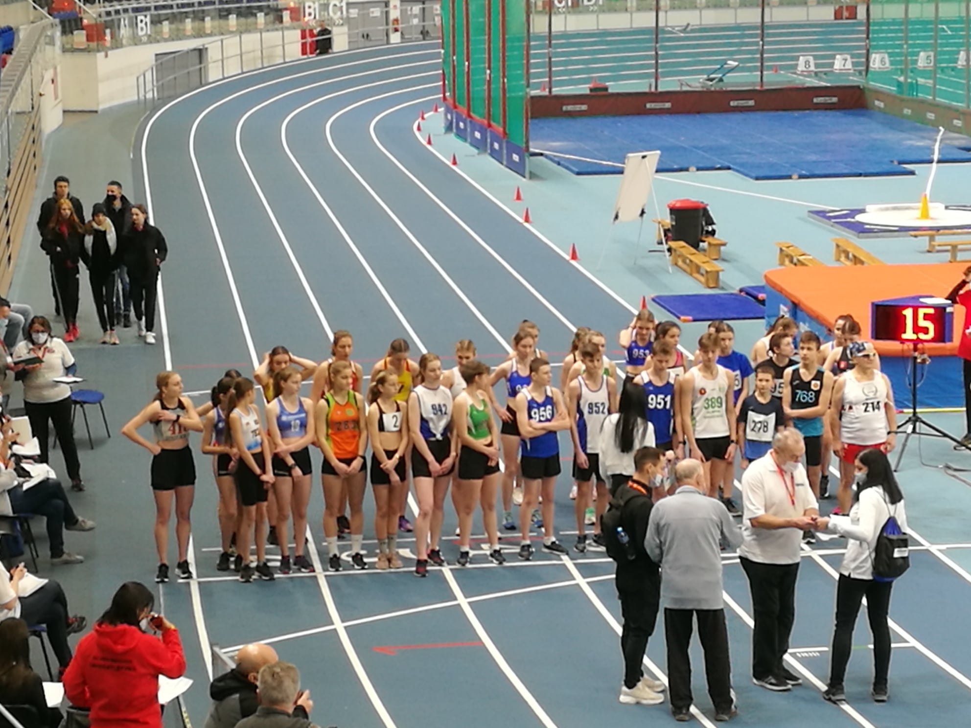 Sportowa hala lekkoatletyczna. Bieżnia. Sportowcy stoją na&nbsp;starcie. Sędziowie przygotowują bieg.