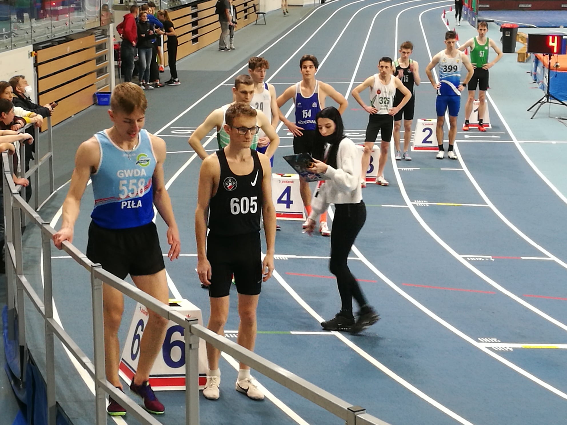 Sportowa hala lekkoatletyczna. Bieżnia. Sportowcy stoją na&nbsp;starcie. Sędziowie przygotowują bieg.