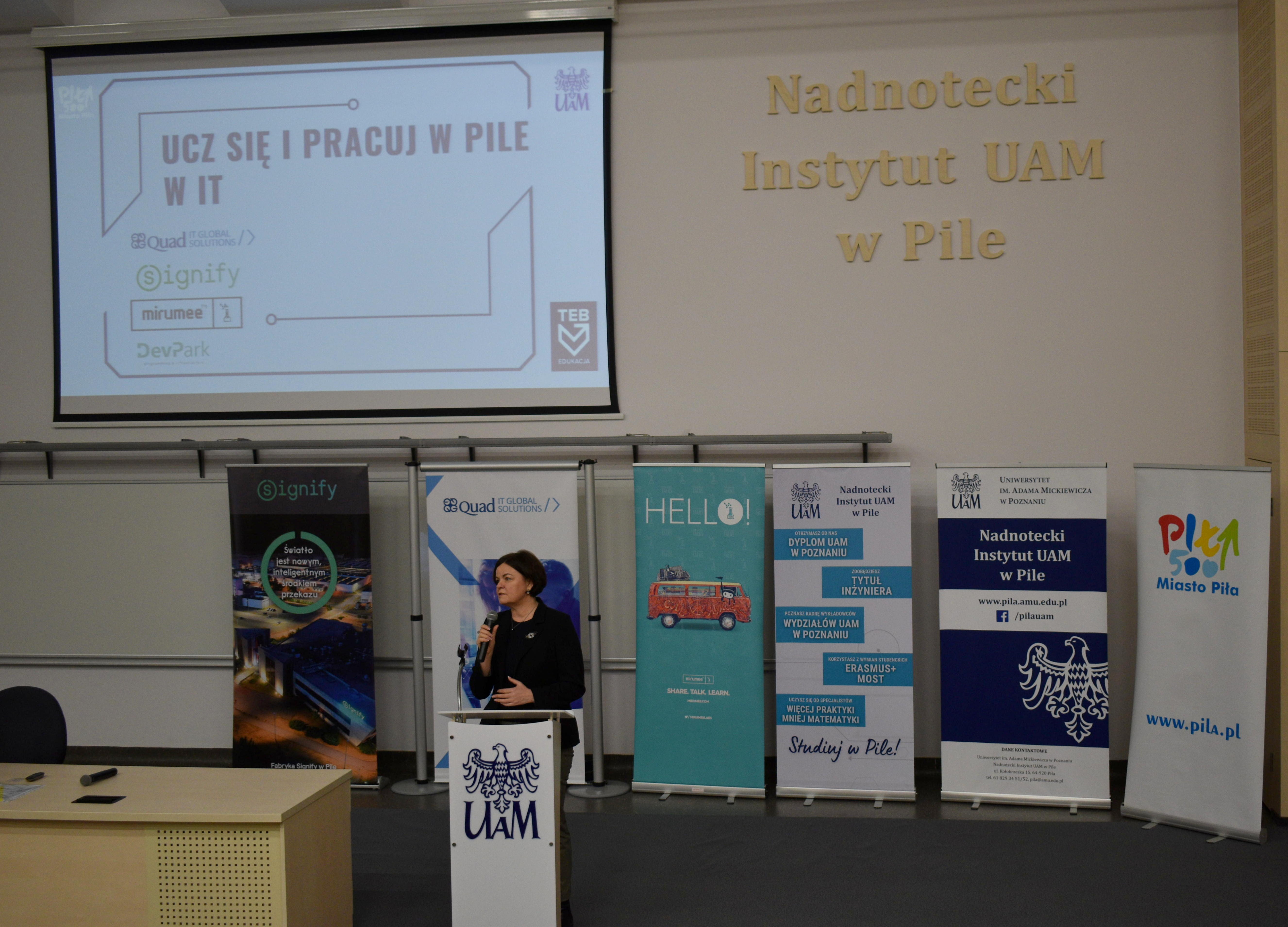 Zdjecie przedstawia kobietę przemawiającą ze sceny w sali wykładowej