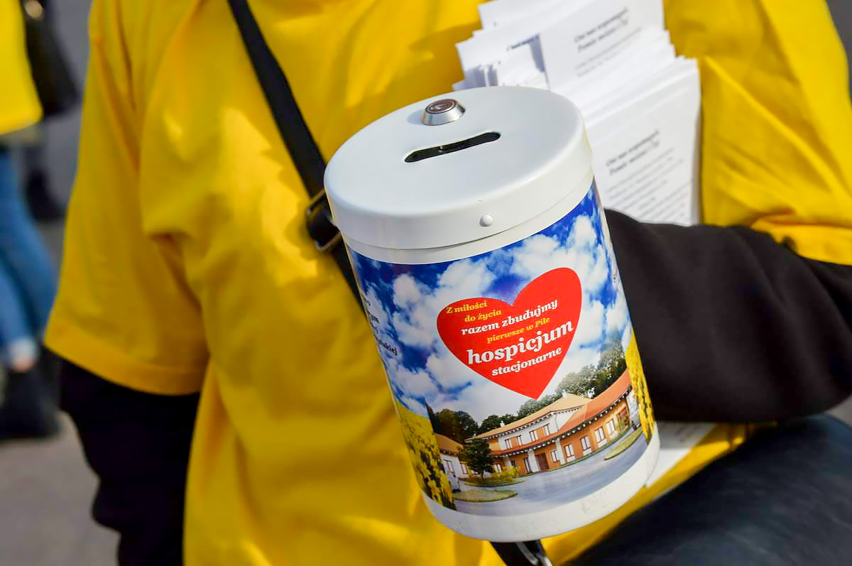 Puszka-skarbonka, zbi&oacute;rka dla pilskiego hospicjum. W tle ż&oacute;łta koszulka wolontariusza, kt&oacute;ry trzyma puszkę. 