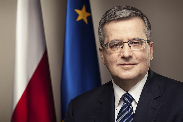 Portret mężczyzny w garniturze na tle flag Polskiej i Unii Europejskiej.