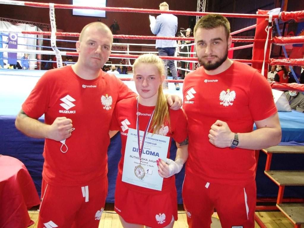 Hala sportowa. Dwaj mężczyźni i&nbsp;kobieta. Zaciśnięte pięści. Dyplom, medal ring bokserski.