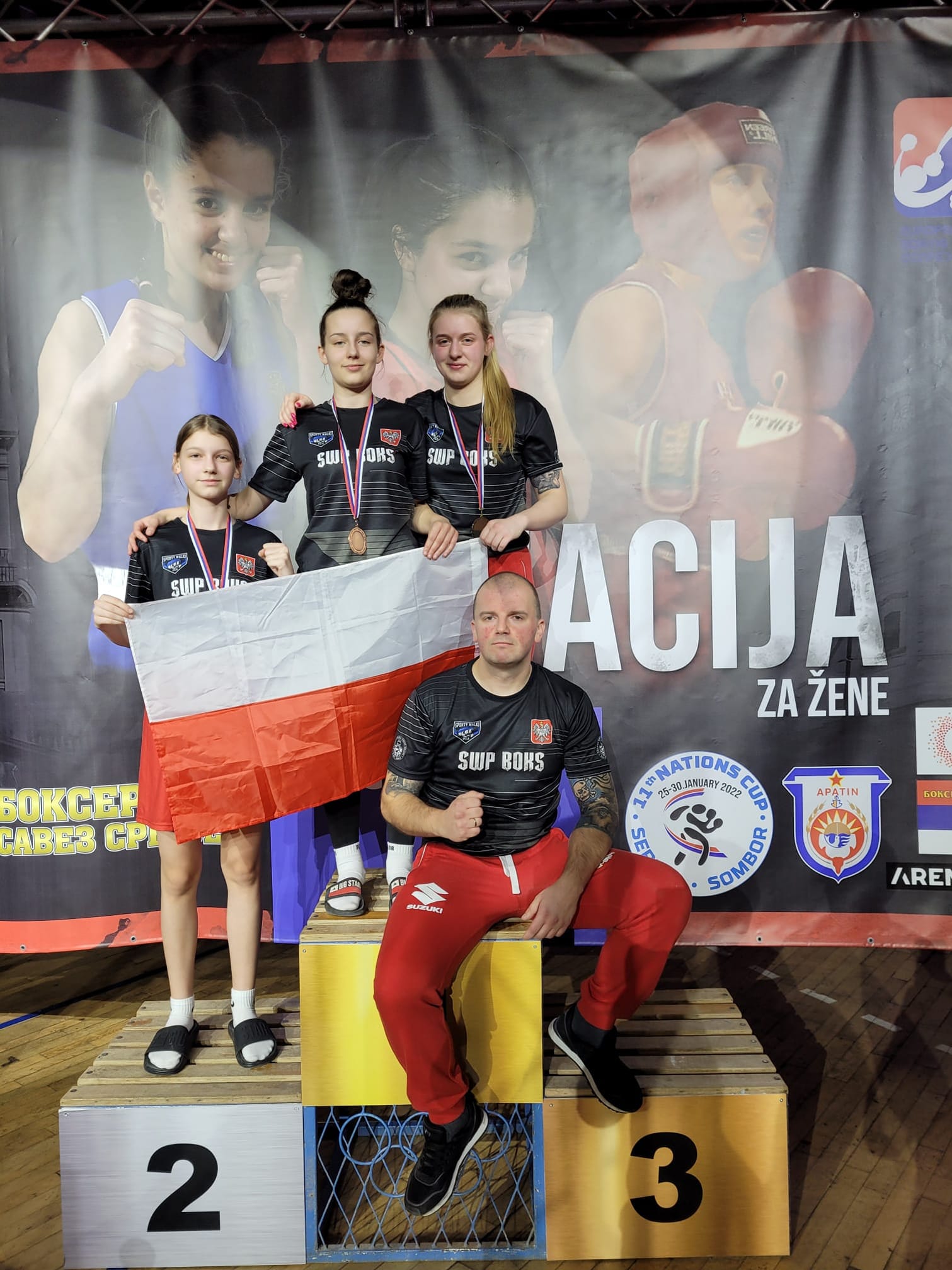 Podium sportowe. Mężczyzna siedzi, trzy kobiety stoją. Polska flaga narodowa.