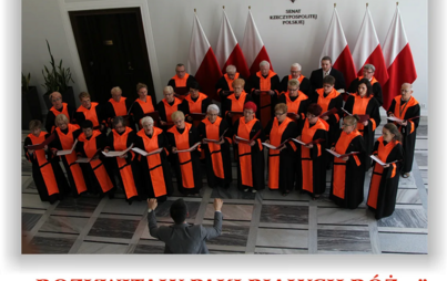 Chór w jednakowych togach, za nimi polskie flagi.