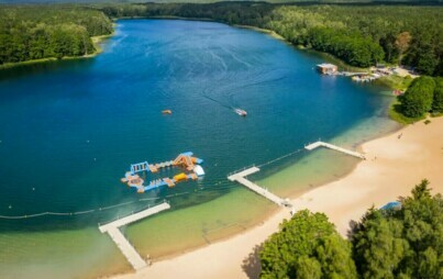 Jezioro i plaża otoczone lasem, wyznaczone kąpielisko z pomostami, dmuchanym torem na wodzie. Zdjęcie z lotu ptaka.