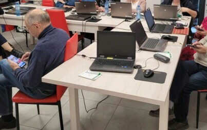 Warsztat dla seniorów, na stole stojące laptopy, wokół siedzący ludzie, trzymający w dłoniach telefony komórkowe