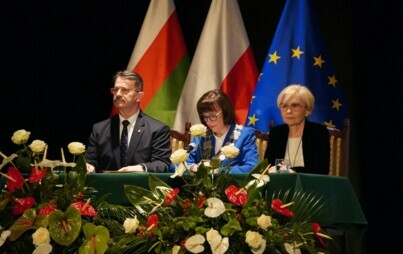 Troje elegancko ubranych ludzi siedzi przy stole, dwie kobiety i mężczyzna. W tle flagi Polski, Piły i Unii Europejskiej, na pierwszym planie przed stołem duże bukiety biało-czerwonych kwiatów.