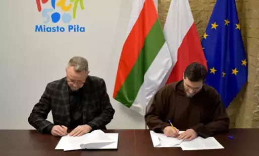 Zastępca prezydenta miasta Piły podpisuje dokumenty razem z przedstawicielem parafii Rzymskokatolickiej pw. św. Antoniego w Pile. Obaj mężczyźni siedzą przy stole, na którym leżą dokumenty i mają w rękach długopisy. W tle widoczne są flagi Polski, Unii Eu