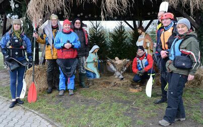 Grupa ludzi w strojach sportowych, niektórzy w koronach, królewskich szatach, mają wiosła. Wszyscy stoją przy drewnianej szopce bożonarodzeniowej.